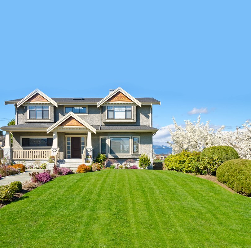 Two-story Craftsman-style house with a covered front porch, gray exterior, and manicured green lawn, surrounded by shrubs and flowering trees under a clear blue sky.