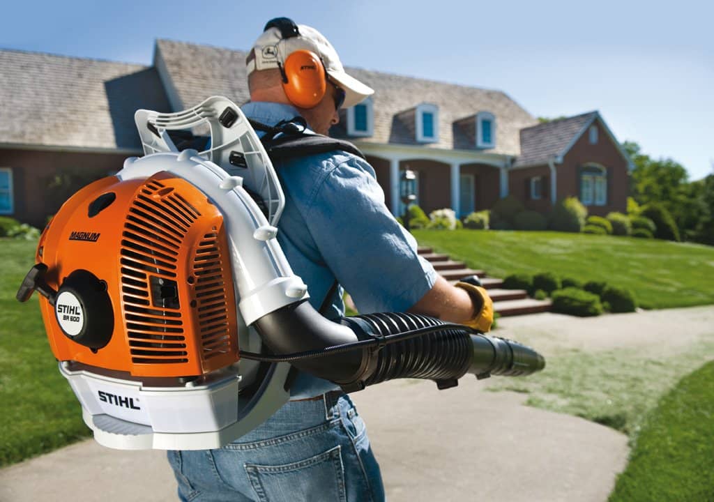 A man operating a backpack leaf blower in front of a large, well-maintained home.