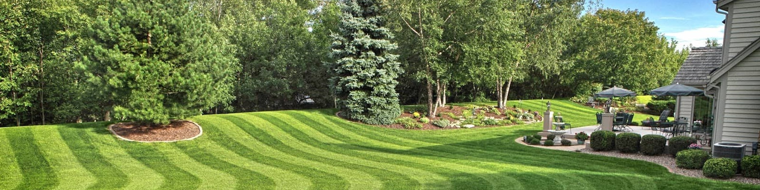 View of a freshly mowed lawn with a home, wooded area, and well-maintained flower bed, and patio.