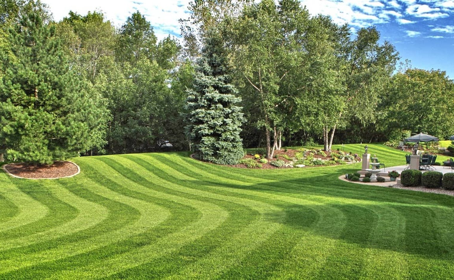 View of a freshly mowed lawn with a home, wooded area, and well-maintained flower bed, and patio.